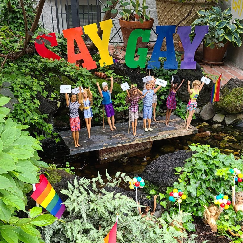 Barbies dressed for pride standing on a rock in a small pond